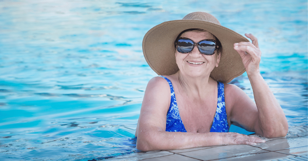 Illustrating modern senior living, an older woman in sun hat and sunglasses smiles from a swimming pool.