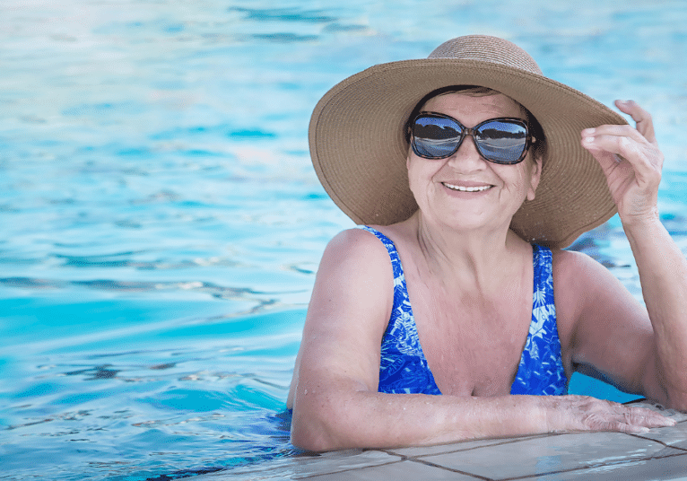 Illustrating modern senior living, an older woman in sun hat and sunglasses smiles from a swimming pool.